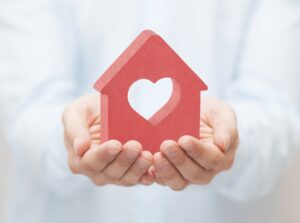 Two hands holding a red wooden house with a heart in the middle.