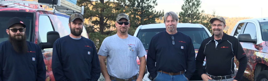 A group of five professional looking HVAC technicians smiling. Behind them are wrapped company vans and pine trees.